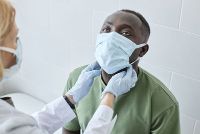 Doctor checking neck of patient wearing protective face mask in clinic