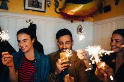 Young multi-ethnic male and female friends holding burning sparklers in restaurant during dinner party