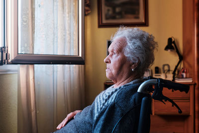 Side view of thoughtful senior disabled female in wheelchair sitting near opened window with curtains in light room at home