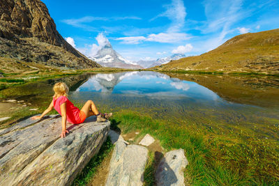 Rear view of woman sitting on rock by lake against sky