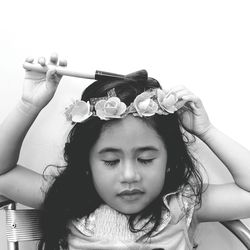 Girl with closed eyes wearing flowers holding brush while sitting on chair against wall