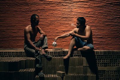 Full length of shirtless man sitting against wall