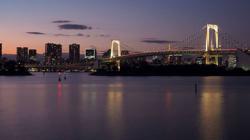 Illuminated bridge over river by buildings against sky at night