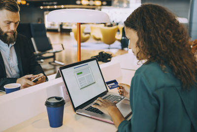 Businesswoman holding credit card while using laptop at table in airport departure area