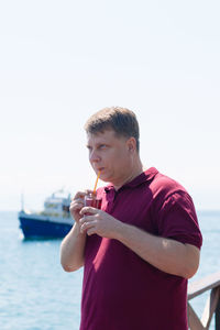 A tall blond man is standing on the pier with a glass of strawberry juice waiting for the ship.