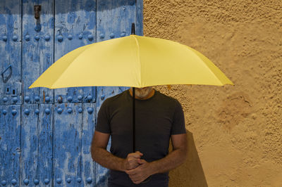 Portrait of adult man with yellow umbrella standing against blue door and yellow wall in summer