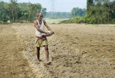 Full length of shirtless farmer spraying seed or fertilizer in plougued farm land