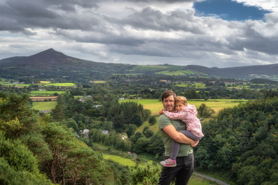 Father holding his daughter in arms with a scenic view on a valley, forest and mountain range