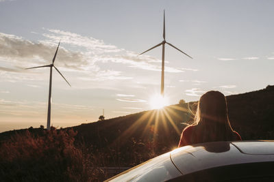 Woman leaning on car looking at wind park at sunset