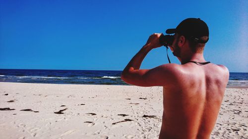 Rear view of shirtless man looking through binoculars at beach