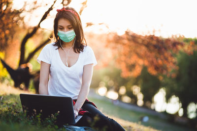 Young woman wearing mask using laptop while sitting at park