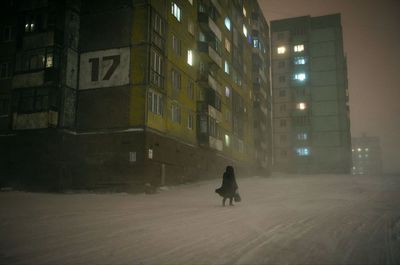 Woman walking on snow covered road against illuminated buildings at night