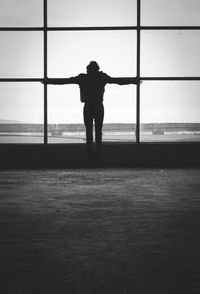 Rear view of man with arms outstretched standing against window in building