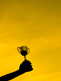 Cropped silhouette hand holding trophy against sky during sunset