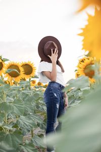 Low angle view of a girl hiding behind a fedora hat in a sunflower field 