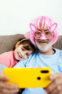 Happy child sitting on couch with grandfather in big pink plastic glasses and pink wig with false ears and making selfie on smartphone in room