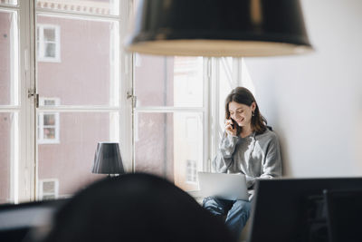 Smiling young female programmer talking over mobile phone while using laptop by window in creative office