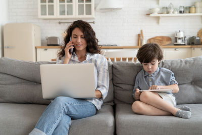 Busy mother talk on smartphone with partners or colleague use laptop computer as son play on tablet