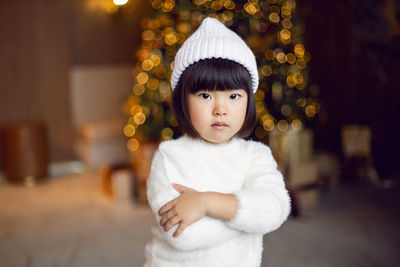 Beautiful korean child girl in in a white knitted sweater and hat stands at the christmas tree 