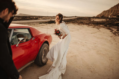 Unrecognizable man in suit standing against red sports car and cheerful bride at sunset in bardenas reales natural park in navarra, spain