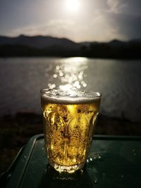 Close-up of beer glass against lake during sunset