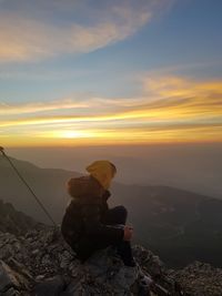 Woman sitting on rock against sky during sunset