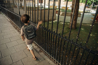 Rear view of boy wearing backpack walking by metal fence