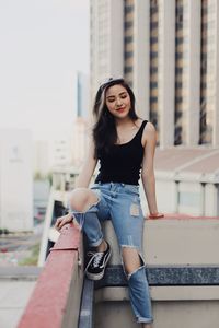 Young woman wearing torn jeans sitting on railing against modern building