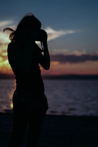 Silhouette woman photographing while standing by sea against sky during sunset