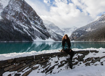 Woman sitting by lake against mountain during winter