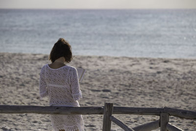 Rear view of woman reading book while leaning on railing at beach