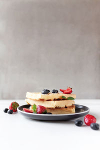 Stack of waffles on a plate on the white table with blueberry, chopped strawberry and mint leaves