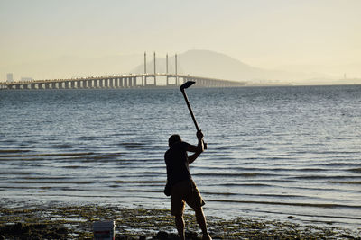 Rear view of man digging with garden hoe at riverbank against bridge during sunset