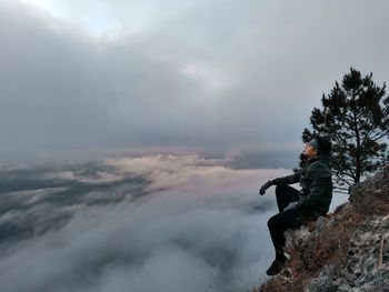 Side view of man sitting on rock against sky
