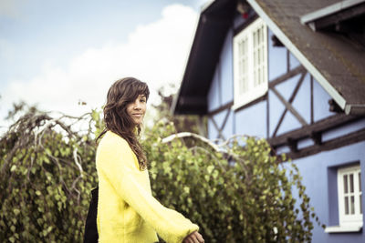 Smiling natural girl in yellow jumper walks by blue traditional house