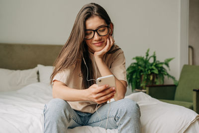 Young smiling female student holding cellphone and scrolling social media feed and chatting at home