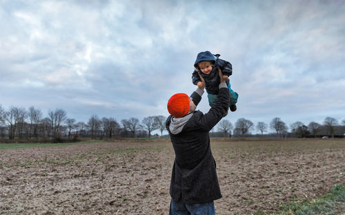 Father picking up son while standing on field against cloudy sky