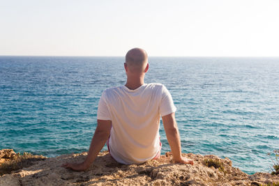 Rear view of man sitting on rock by sea against clear sky