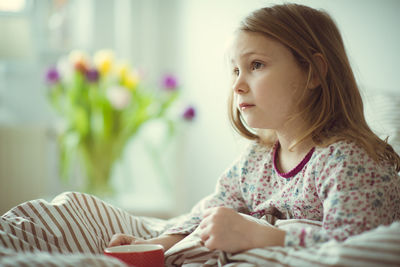 Cute girl holding cup looking away while sitting on bed