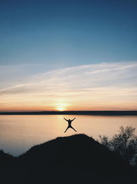 Silhouette person jumping against lake during sunset