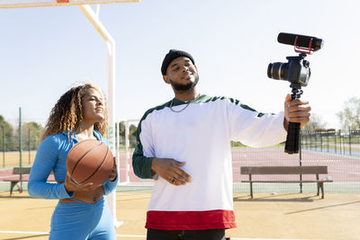 Male holding tripod with camera while vlogging with female friend on sports court