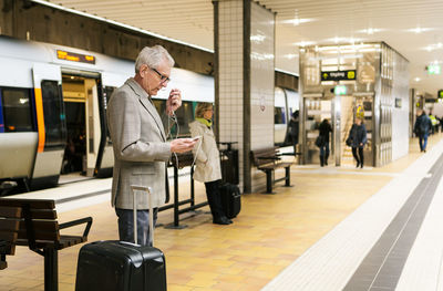Senior male commuter using smart phone while standing by luggage at subway station