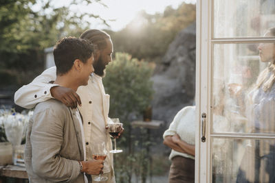 Man with arm around male friend holding wineglass while standing during dinner party at cafe