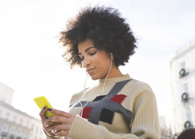 Young woman wearing in-ear headphones using mobile phone while standing outdoors