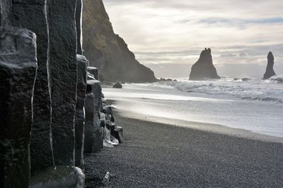 Rock formations by black beach against sky
