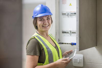 Portrait of smiling female electrician with tablet at fusebox