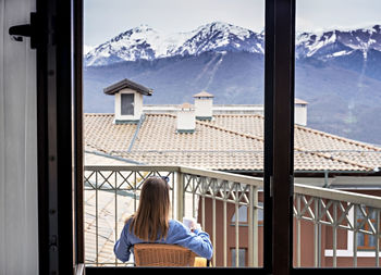 Rear view of blond woman in blue sitting with mug of drink on terrace and looking at mountain view 