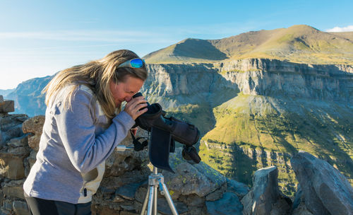 Blonde woman at ordesa national park looking thought a telescope on a tripod some details of the park. behind the woman there are some peaks and rocks of the panoramic view. horizontal photo