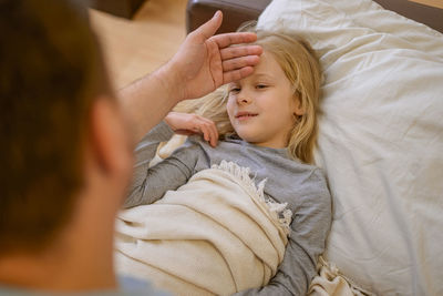 Dad touches the forehead of his sick child, who is lying on the couch, covered with a white blanket