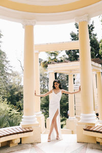 A beautiful brunette lady in an elegant wedding dress poses among the columns in the old city park